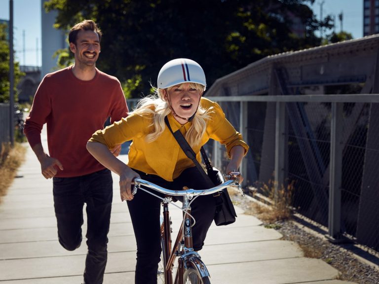 Junge Frau mit Helm fährt Fahrrad, während ein Mann lachend hinterherläuft.